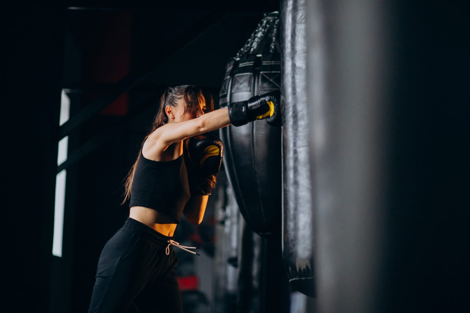 A woman in black training gear punches a heavy bag in a dark gym.