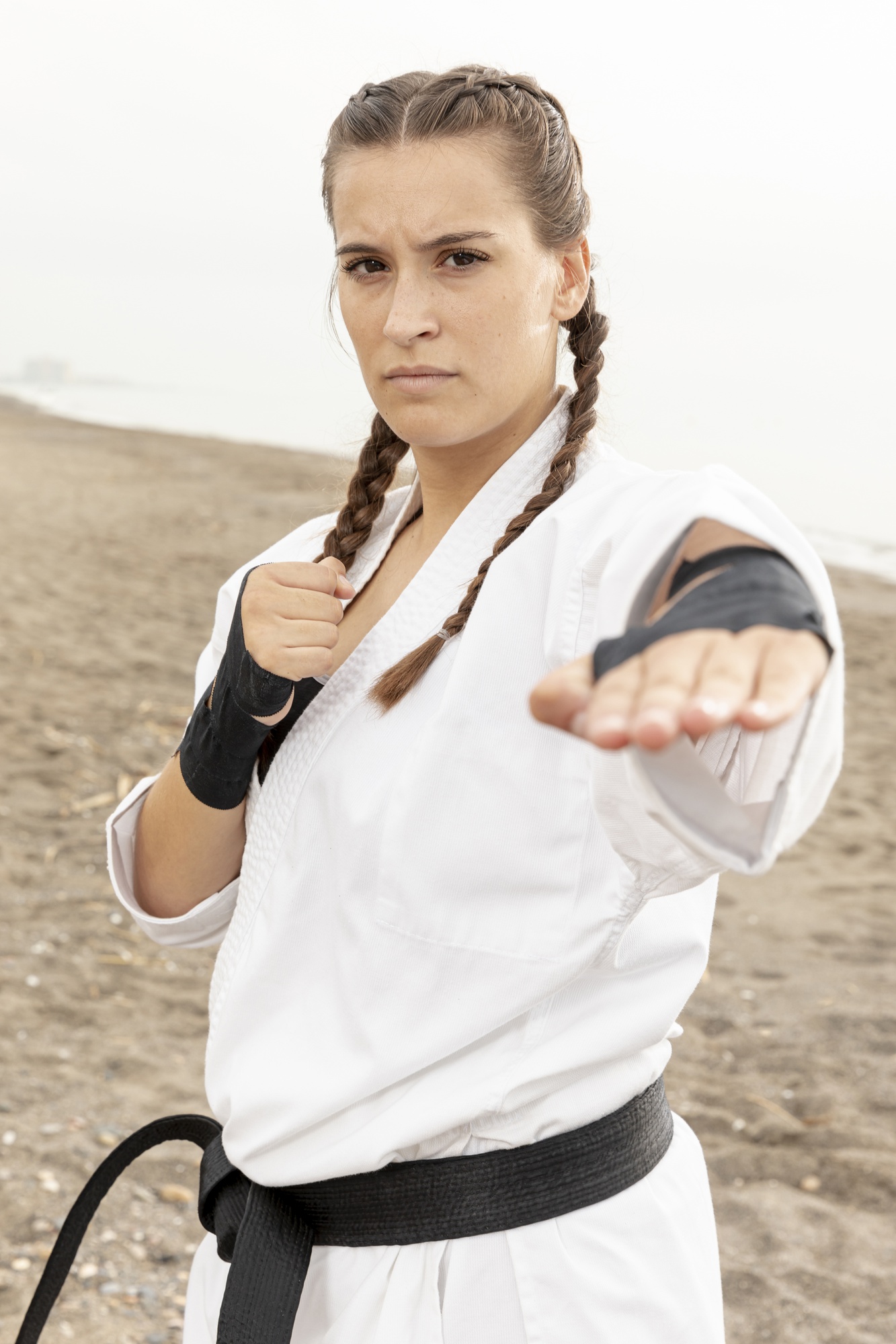 A woman in a white gi and black belt extends an open-hand block on a foggy beach.