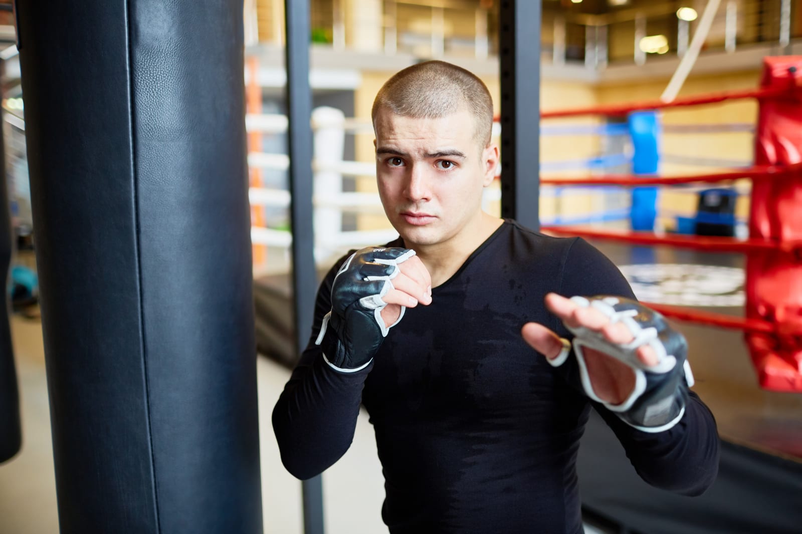 A man in a black rash guard and grappling gloves holds a guard beside a heavy bag and ring.