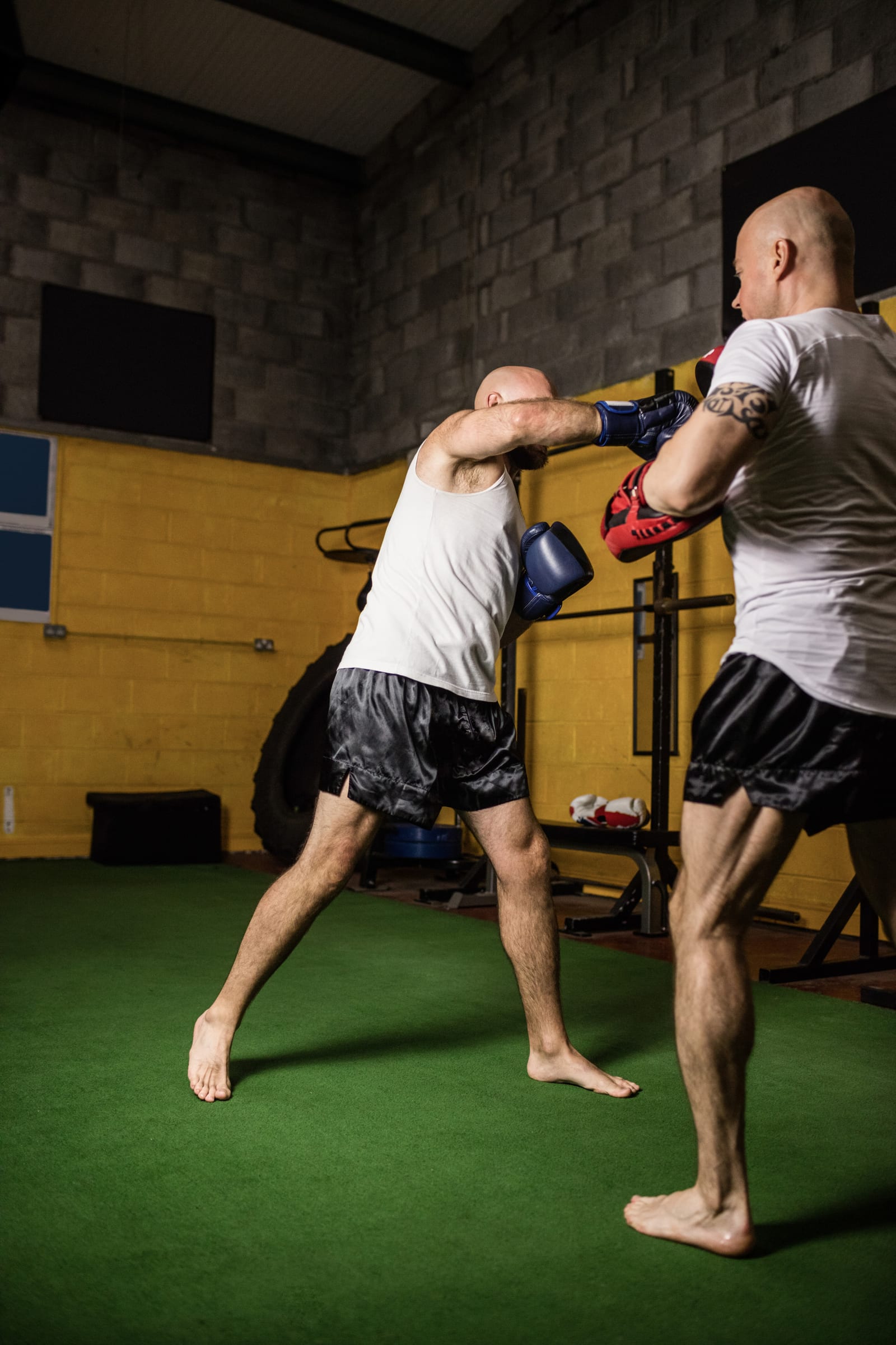 Two men in shorts spar with high kicks on the green floor of a training hall.
