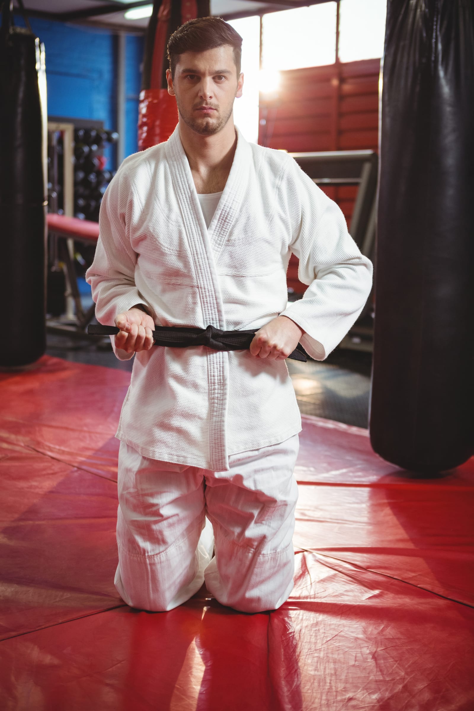 A man in a white gi kneels on a red mat tying his black belt beside a heavy bag.
