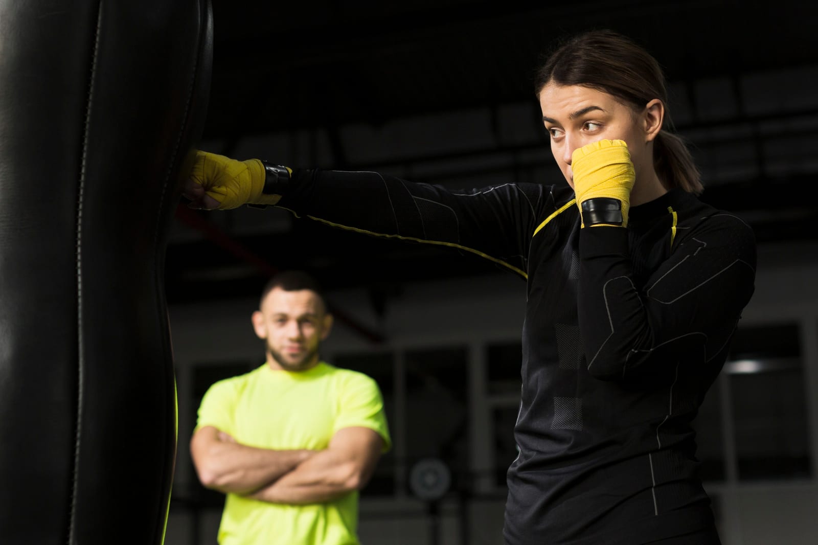 A woman with yellow hand wraps punches a heavy bag while a coach watches from behind.